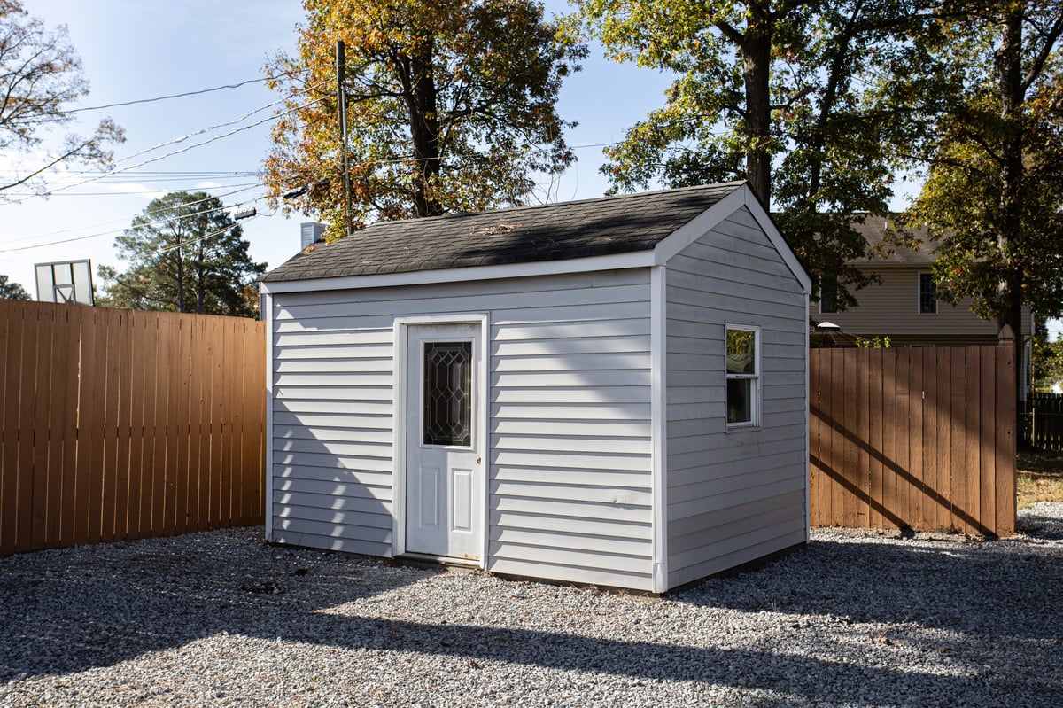 A quaint wooden shed with white siding stands on gravel, enclosed in a fenced yard, surrounded by mature trees